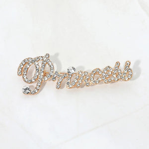 Gold-tone “Princess” brooch with clear crystals covering flowing, connected script lettering and a large looping capital P, measuring approximately 2.8 inches wide by 1.1 inches tall, displayed on a white background.