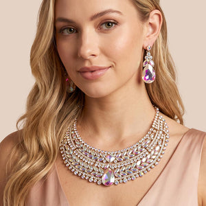 AB iridescent crystal statement necklace set on gold-tone metal, featuring a broad, layered bib of marquise and round stones with a large teardrop centerpiece and matching teardrop earrings reflecting rainbow colors. Necklace measures 17 inches plus a 2-inch extender, with 2.25-inch decor; earrings are 2.2 inches long. Worn by a woman in a blush sleeveless outfit against a warm beige background.