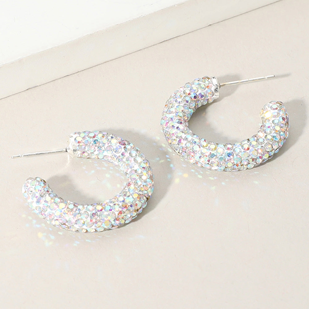 A pair of silver hoop-style earrings covered in iridescent, rainbow-reflecting rhinestones, displayed on a light beige surface.