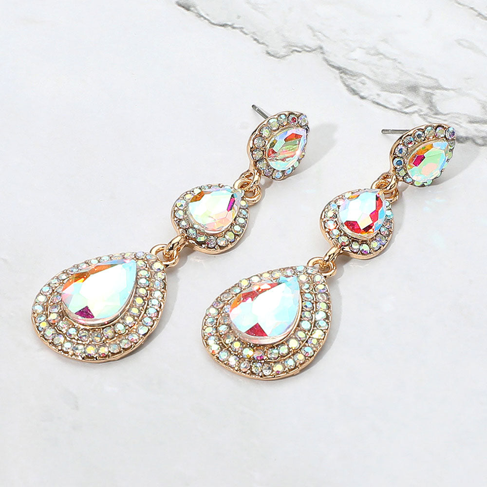 A pair of gold-tone drop earrings with three cascading teardrop-shaped AB gemstones, each surrounded by a halo of small clear rhinestones, displayed on a light textured background.