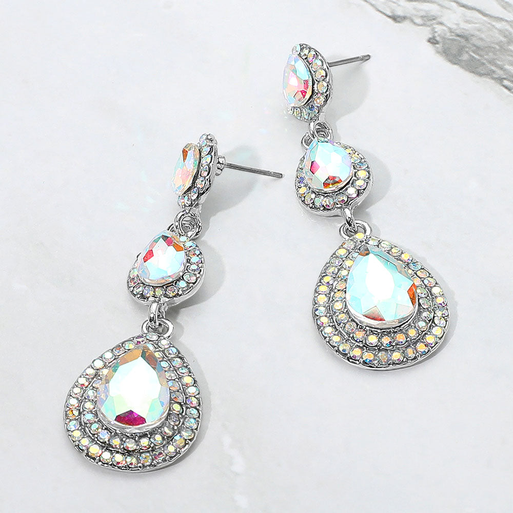 A pair of silver-tone drop earrings with three cascading teardrop-shaped AB gemstones, each surrounded by a halo of small clear rhinestones, displayed on a light textured background.
