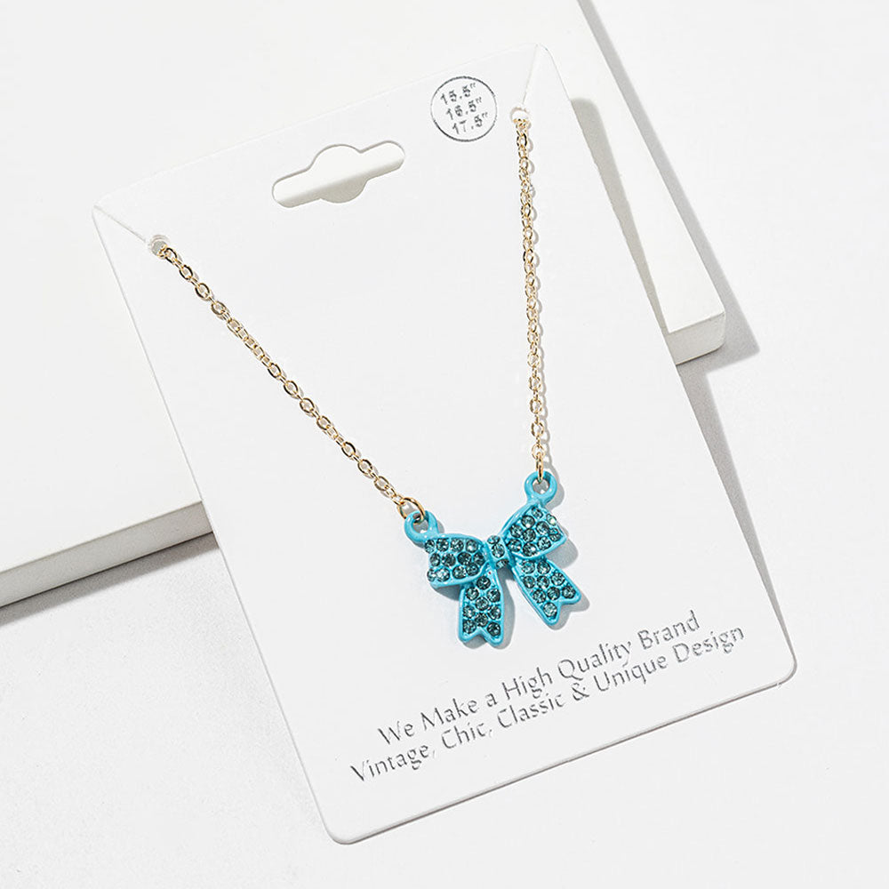 Bow pendant necklace with aqua crystals in an aqua setting on a gold-tone chain, measuring 15.5 inches with a 2-inch extender. The bow measures 0.75 inch wide by 0.75 inch long, with two rounded loops, a center knot, and two ribbon tails covered with small round crystals, displayed on a white jewelry card.