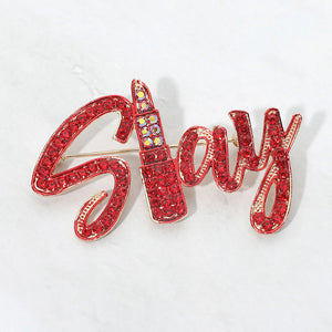 Gold-tone “Slay” brooch with red crystals covering flowing, connected script lettering, measuring approximately 2.6 inches wide by 1.5 inches tall on a white background.
