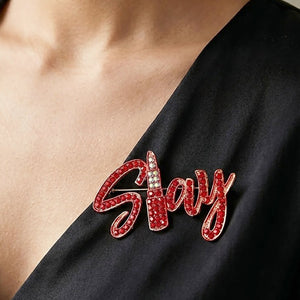 Gold-tone “Slay” brooch with red crystals covering flowing, connected script lettering, measuring approximately 2.6 inches wide by 1.5 inches tall shown here on a black lapel