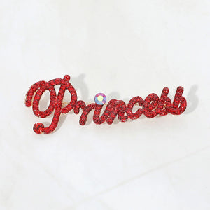 Gold-tone “Princess” brooch with red crystals covering flowing, connected script lettering and a large looping capital P, measuring approximately 2.8 inches wide by 1.1 inches tall, displayed on a white background.