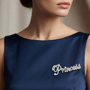 Silver-tone “Princess” brooch with clear crystals covering flowing, connected script lettering and a large looping capital P, measuring approximately 2.8 inches wide by 1.1 inches tall, displayed on a model's navy dress.