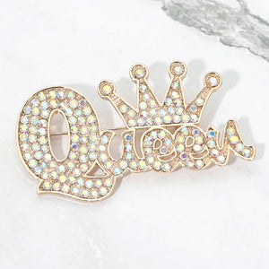 AB iridescent crystal “Queen” brooch on gold-tone metal, featuring connected script lettering with a large looping capital Q and a five-point crown above the smaller letters. Measures approximately 2.75 inches wide by 1.5 inches tall, displayed on a white marble surface.