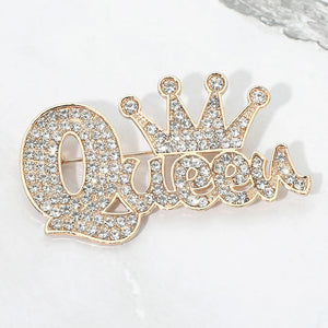 Clear crystal “Queen” brooch on gold-tone metal, featuring connected script lettering with a large looping capital Q and a five-point crown above the smaller letters. Measures approximately 2.75 inches wide by 1.5 inches tall, displayed on a white marble surface.