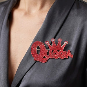 Red crystal “Queen” brooch on silver-tone metal, featuring connected script lettering with a large looping capital Q and a five-point crown above the smaller letters. Measures approximately 2.75 inches wide by 1.5 inches tall. Pinned to the lapel of a black blazer in a close-up photograph.