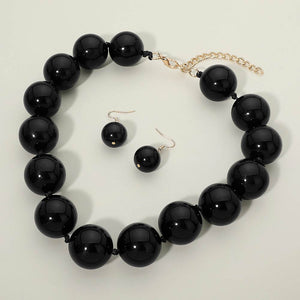 Black chunky ball beaded statement necklace set with oversized round beads, matching drop earrings, displayed on a light cream background.