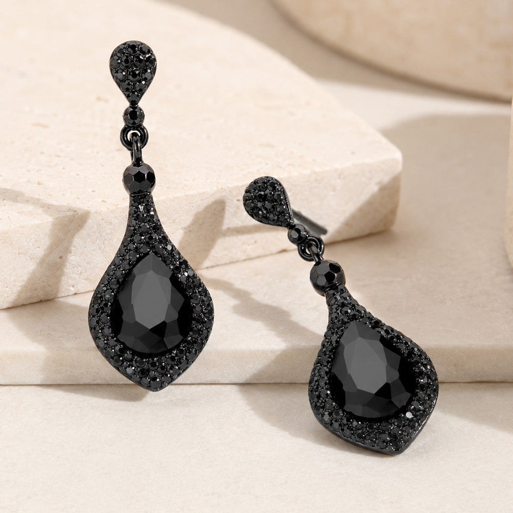 A pair of black drop earrings adorned with black crystals, featuring a large teardrop crystal surrounded by smaller black crystals, displayed on a neutral beige background.
