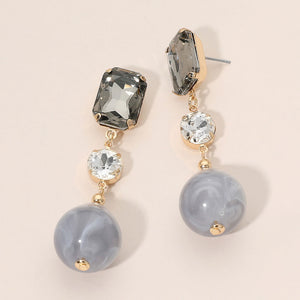 Gray faceted crystal and ball drop statement earrings on gold with rectangular, round, and spherical gray stones, displayed on a light beige background.
