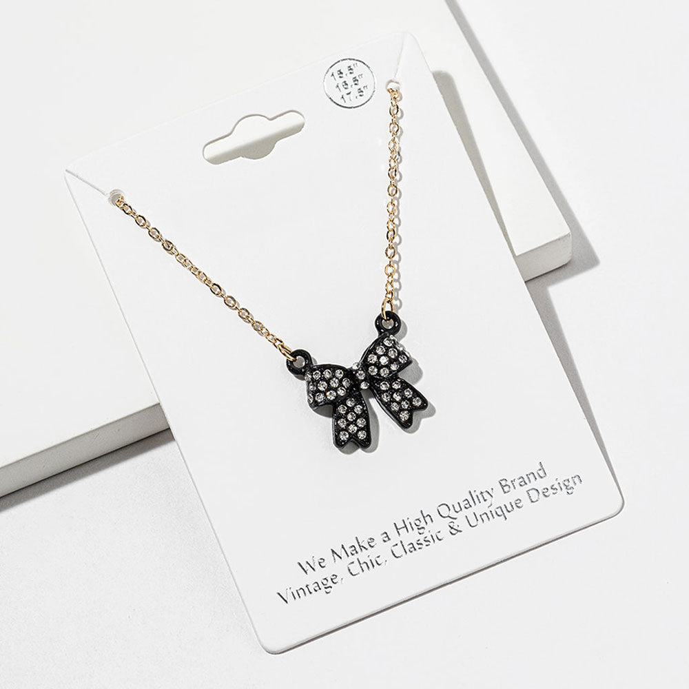 Bow pendant necklace with clear crystals set inside a black bow on a gold-tone chain, measuring 15.5 inches with a 2-inch extender. The bow measures 0.75 inch wide by 0.75 inch long, with two rounded loops, a center knot, and two ribbon tails outlined in black, displayed on a white jewelry card.
