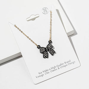 Bow pendant necklace with clear crystals set inside a black bow on a gold-tone chain, measuring 15.5 inches with a 2-inch extender. The bow measures 0.75 inch wide by 0.75 inch long, with two rounded loops, a center knot, and two ribbon tails outlined in black, displayed on a white jewelry card.
