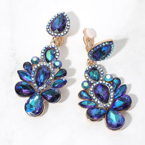Gold-tone chandelier clip-on earrings with blue AB teardrop and oval faceted crystals surrounded by smaller clear rhinestones, displayed on a white marble surface.