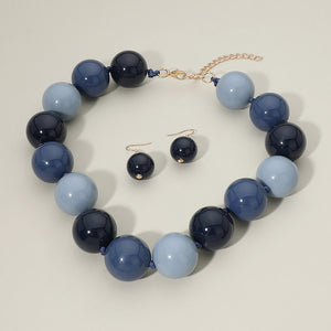 Navy, blue, and light blue chunky ball beaded statement necklace set with oversized round beads and matching earrings, displayed on a beige background.