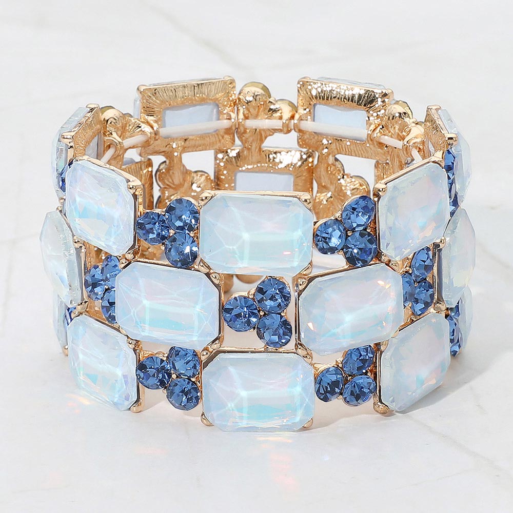 A gold-tone stretch bracelet featuring rectangular light blue opal-like crystals and round darker blue faceted rhinestones, displayed on a light marble background.
