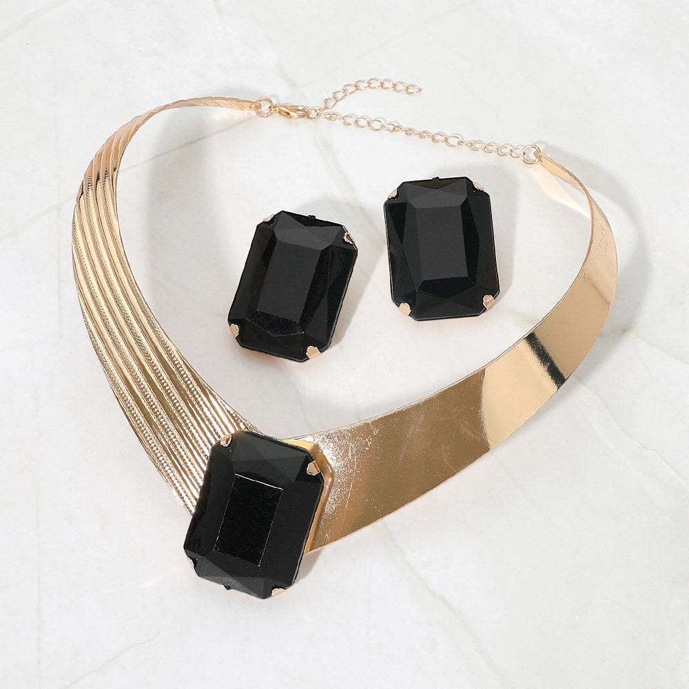 Bold black crystal statement choker necklace set on gold with a large rectangular center stone, structured metallic band, and matching earrings, displayed on a white marble background.