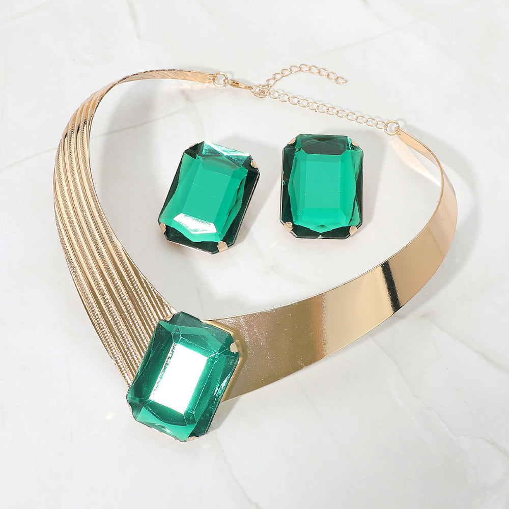 Bold Emerald Green crystal statement choker necklace set on gold with a large rectangular center stone, structured metallic band, and matching earrings, displayed on a white marble background.