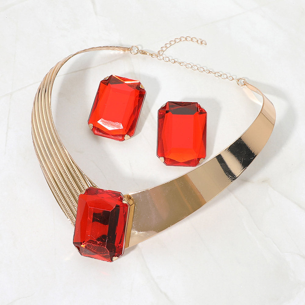 Bold red crystal statement choker necklace set on gold with a large rectangular center stone, structured metallic band, and matching earrings, displayed on a white marble background.