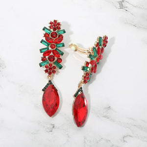 A pair of gold-tone clip-on dangle earrings decorated with red marquise and round crystals, accented by green baguette and round crystals, displayed on a white marble surface.