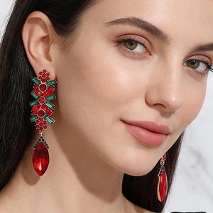 A close-up of a woman wearing a pair of ornate clip on earrings with red floral clusters, green leaf-like accents, and large red teardrop stones.