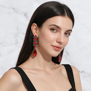 A woman with long dark hair models a pair of long, ornate red and green teardrop clip on earrings against a light gray marble background.
