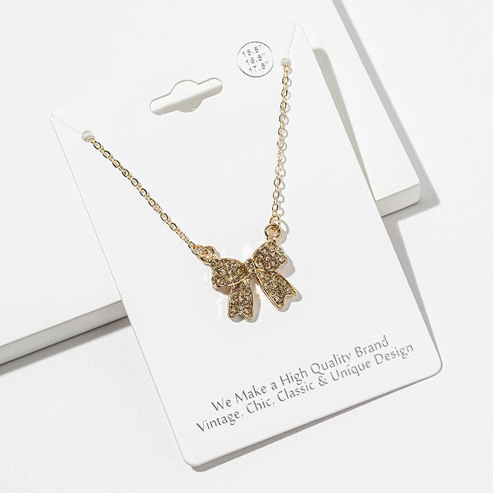 Bow pendant necklace with champagne gold crystals on a gold-tone setting and chain, measuring 15.5 inches with a 2-inch extender. The bow measures 0.75 inch wide by 0.75 inch long, with two rounded loops, a center knot, and two ribbon tails covered with small round crystals, displayed on a white jewelry card.