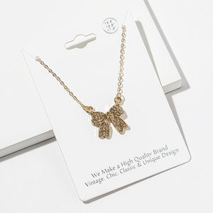 Bow pendant necklace with champagne gold crystals on a gold-tone setting and chain, measuring 15.5 inches with a 2-inch extender. The bow measures 0.75 inch wide by 0.75 inch long, with two rounded loops, a center knot, and two ribbon tails covered with small round crystals, displayed on a white jewelry card.