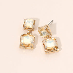 Champagne gold-colored crystal double-square drop earrings on gold-tone metal, each featuring a smaller faceted square stone connected to a larger square stone below. Each earring measures approximately 0.4 inches wide by 0.9 inches long. Displayed on a pale peach surface.