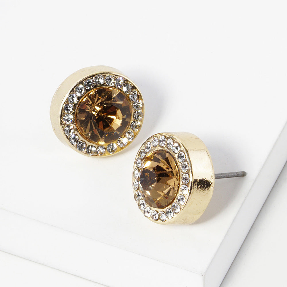 A pair of gold stud earrings with an oval Champagne Gold Stone faceted center stone surrounded by a halo of small clear rhinestones, displayed on a clean white surface.