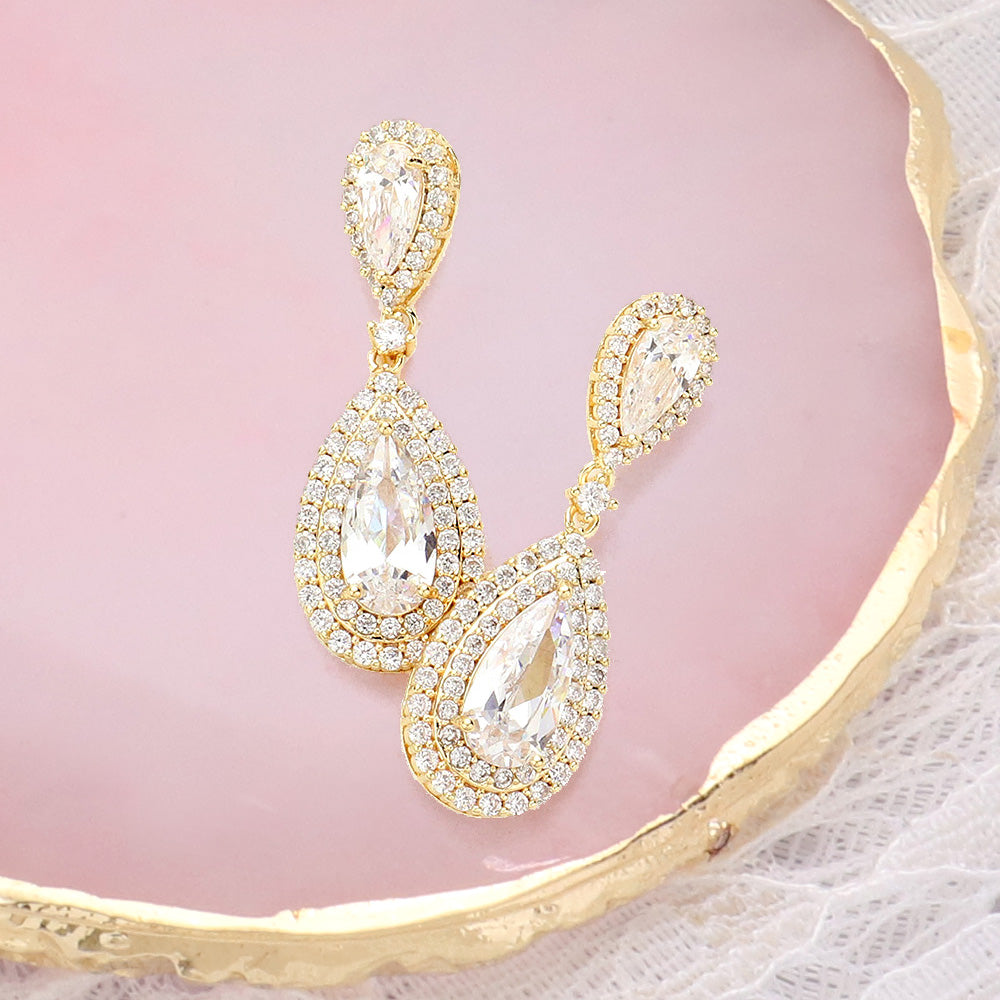 Clear crystal double teardrop drop earrings in a gold finish featuring pear-shaped crystal centers surrounded by double teardrop halos, displayed on a pink jewelry background.