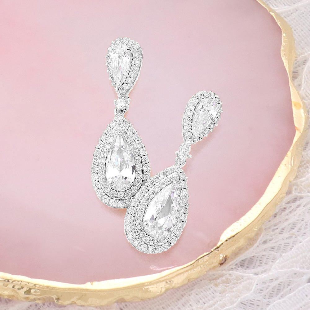 Clear crystal double teardrop drop earrings in a silver finish featuring pear-shaped crystal centers surrounded by double teardrop halos, displayed on a pink jewelry background.