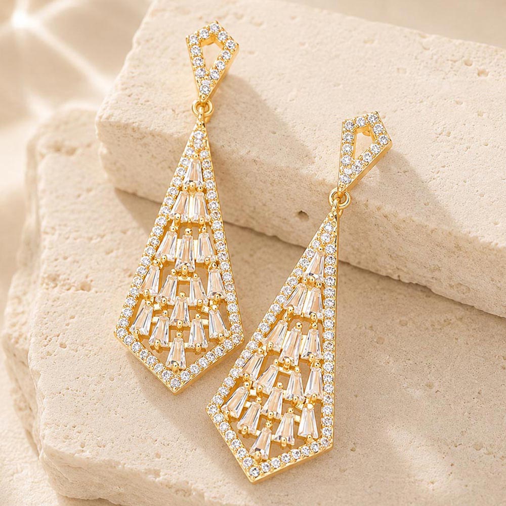 Clear crystal geometric baguette drop earrings in a gold finish featuring baguette-cut clear crystals arranged in an open geometric drop design, displayed on a beige stone background.