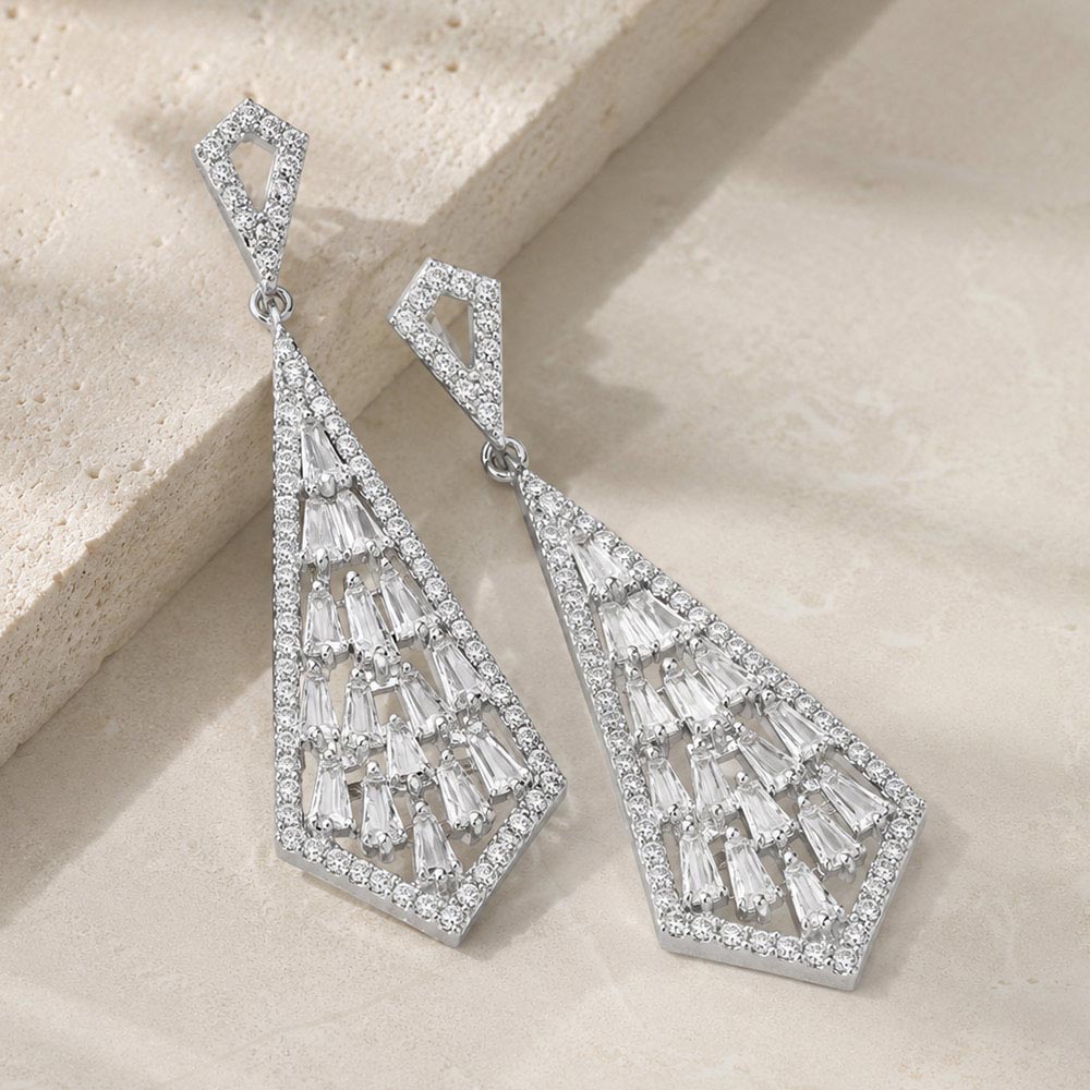 Clear crystal geometric baguette drop earrings in a silver finish featuring baguette-cut crystals arranged in an open geometric drop design, displayed on a beige stone background.