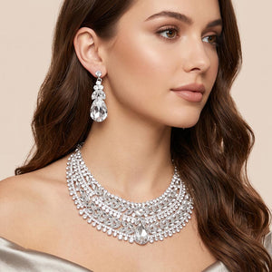 Clear crystal statement necklace set on silver-tone metal, featuring a broad, layered bib of marquise and round stones with a large teardrop centerpiece and matching teardrop earrings. Necklace measures 17 inches plus a 2-inch extender, with 2.25-inch decor; earrings are 2.2 inches long. Worn by a woman in an off-the-shoulder champagne satin outfit against a warm beige background.