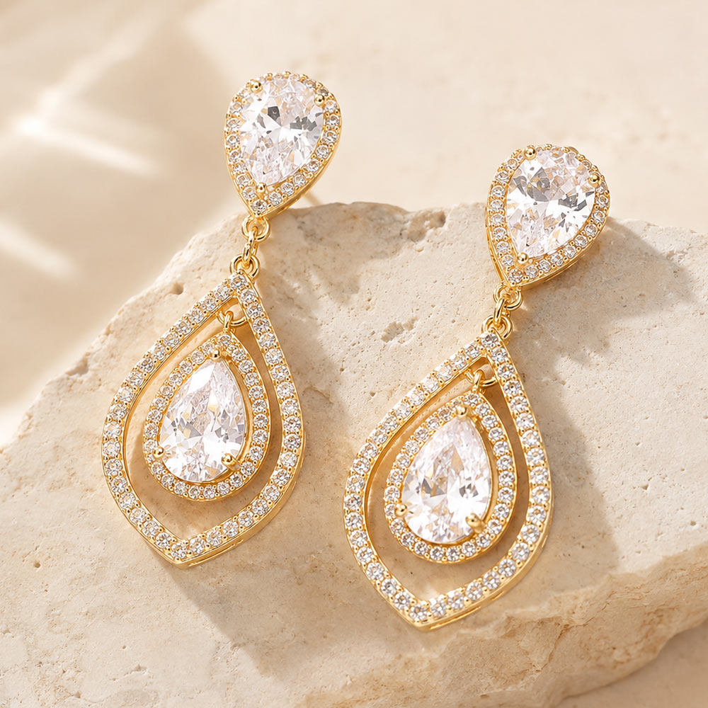 Clear crystal open teardrop drop earrings in a gold finish featuring pear-shaped crystal studs and layered open teardrop frames with clear stones, displayed on a beige stone background.