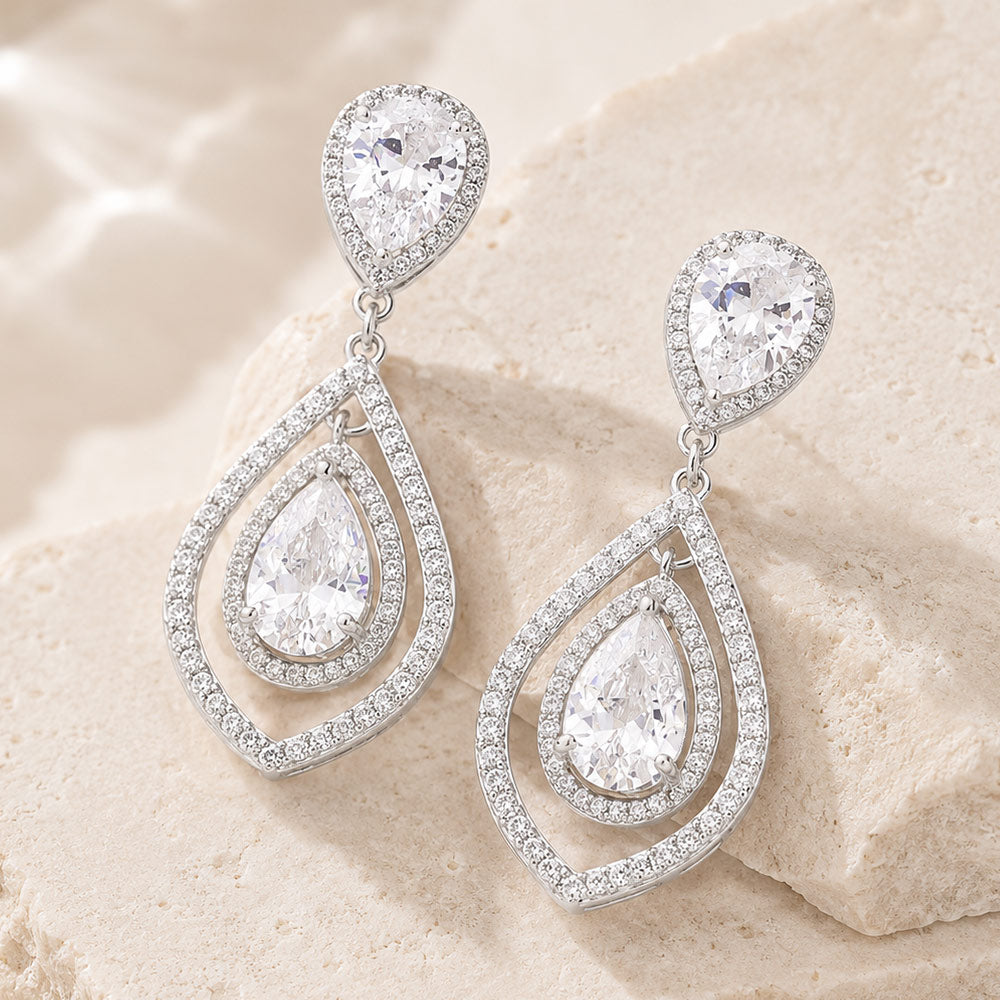Clear crystal open teardrop drop earrings in a silver finish featuring pear-shaped crystal studs and layered open teardrop frames with clear stones, displayed on a beige stone background.