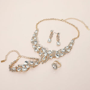 Clear crystal 4-piece jewelry set on gold with statement necklace, matching bracelet, drop earrings and adjustable ring arranged on a light beige background.