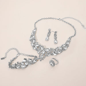 Clear crystal 4-piece jewelry set on silver with statement necklace, matching bracelet, drop earrings and adjustable ring arranged on a light beige background.