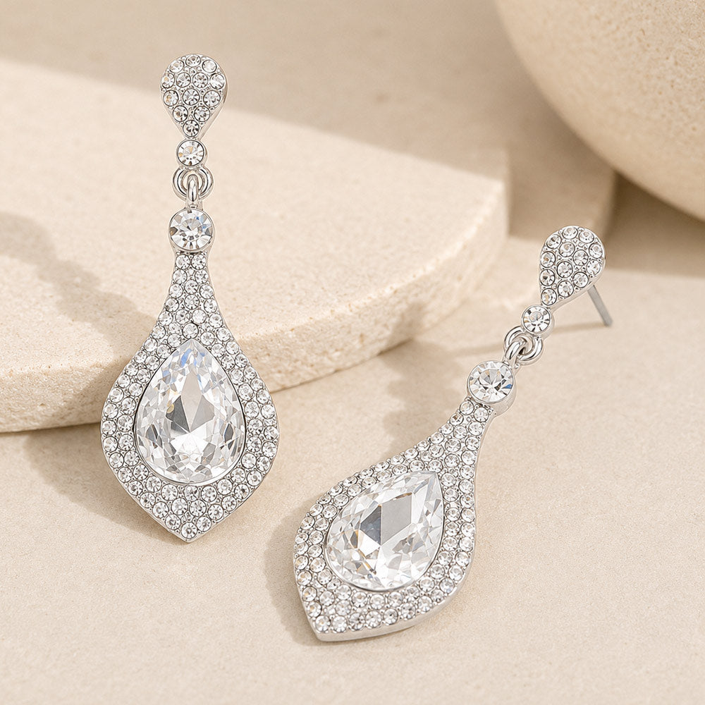 Clear crystal teardrop drop earrings on silver featuring a large clear teardrop crystal surrounded by smaller clear crystals, displayed on a neutral beige background.