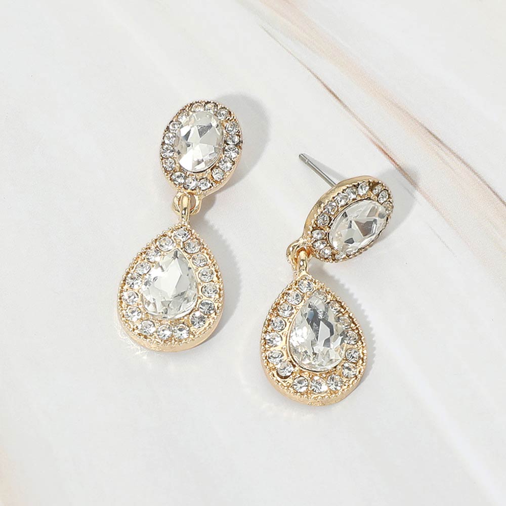 A pair of gold-tone teardrop dangle earrings with clear crystal center stones surrounded by smaller clear rhinestones, displayed on a white marble surface.