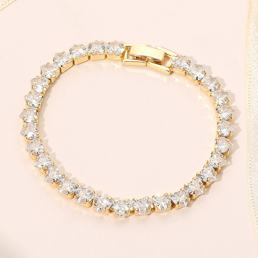 Clear crystal heart tennis bracelet in a gold finish featuring a continuous row of heart-shaped clear crystals with a fold-over clasp, displayed on a light jewelry background.