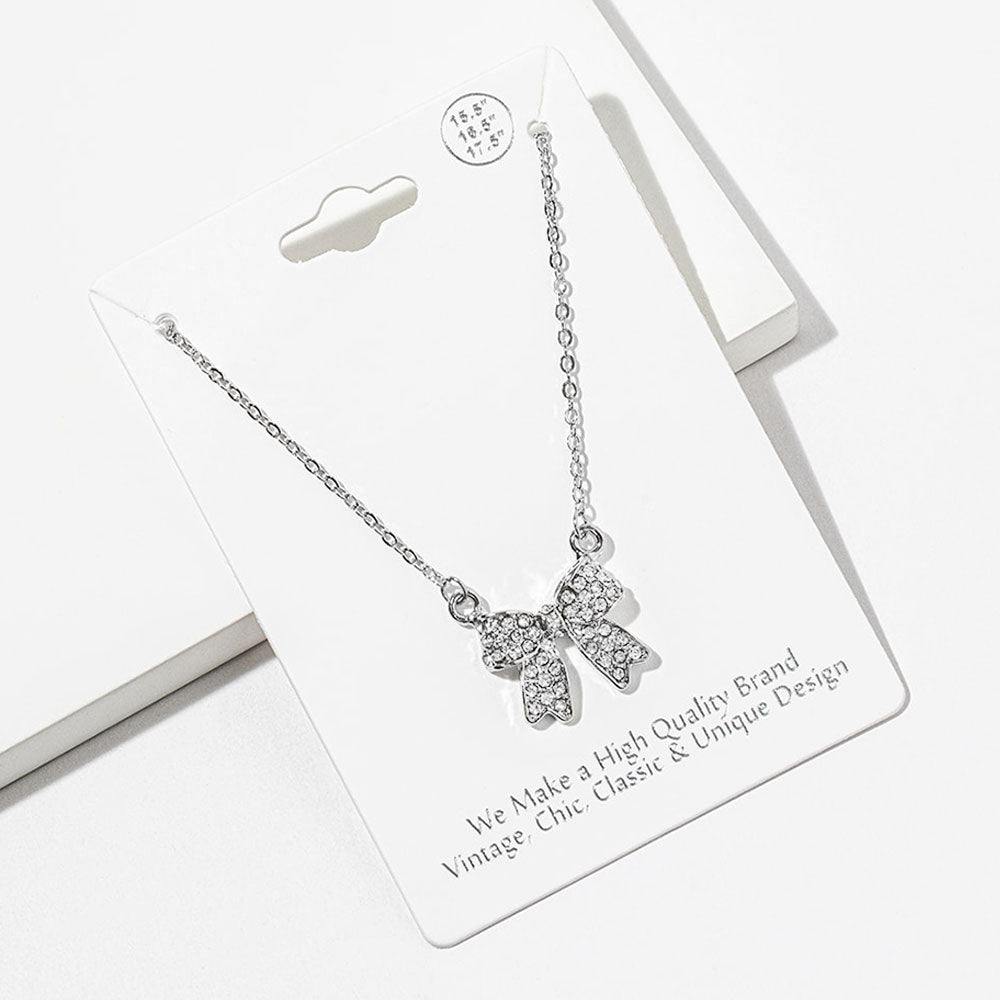 Bow pendant necklace with clear crystals in a silver-tone setting on a silver-tone chain, measuring 15.5 inches with a 2-inch extender. The bow measures 0.75 inch wide by 0.75 inch long, with two rounded loops, a center knot, and two ribbon tails covered with small round crystals, displayed on a white jewelry card.