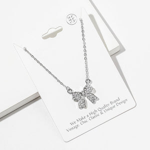 Bow pendant necklace with clear crystals in a silver-tone setting on a silver-tone chain, measuring 15.5 inches with a 2-inch extender. The bow measures 0.75 inch wide by 0.75 inch long, with two rounded loops, a center knot, and two ribbon tails covered with small round crystals, displayed on a white jewelry card.