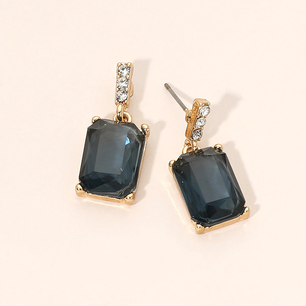 Gold-tone dangle earrings featuring large rectangular navy blue crystals with a short row of small clear crystals above each stone, displayed on a light beige background.