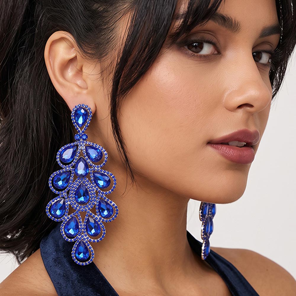 A close-up of a woman wearing a extra long, ornate sapphire royal blue teardrop dangle earring . The earring features multiple layers of teardrop-shaped blue gems surrounded by silver-toned rhinestone borders.