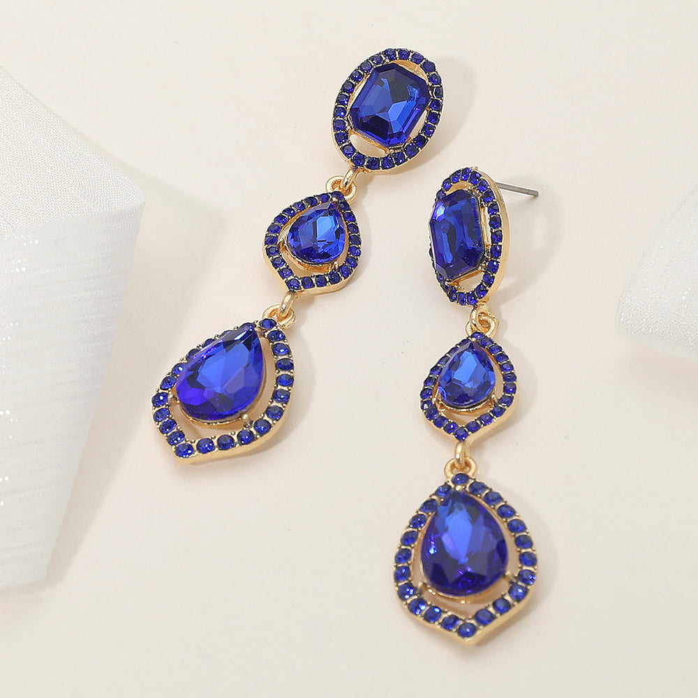Pair of gold-tone drop earrings featuring large sapphire royal blue oval and pear-shaped crystals surrounded by small clear stones on a light gray background.