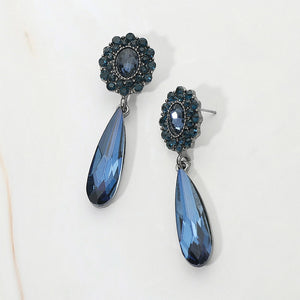 A pair of drop earrings with navy teardrop-shaped crystals and navy floral headpieces, backed in gun metal, displayed on a light off-white background.