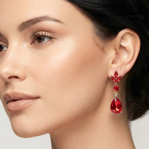 A close-up of a woman's ear wearing a red teardrop dangle earring with gold-toned metal detailing and small red stones above the drop.